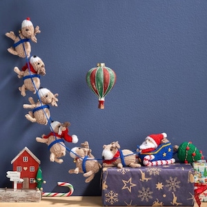 Puede incluir: Una escena navideña caprichosa con renos de punto tirando de un trineo de Papá Noel. Un globo aerostático rojo y verde flota arriba. También hay una pequeña casa roja, un bastón de caramelo y un árbol de Navidad. La escena está ambientada sobre un fondo azul.