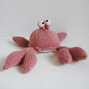 May include: A knitted crab toy in a dusty rose colour with a rounded body, two protruding eyes on stalks with black beads for pupils, and multiple segmented legs and claws extending from the sides, all crafted with a visible knit texture.