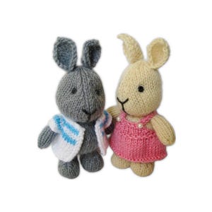 Puede incluir: Dos muñecos de conejos tejidos. Uno es gris y lleva una chaqueta blanca con rayas azules. El otro es beige y lleva un vestido rosa con detalles de perlas. Ambos tienen ojos y nariz negros.