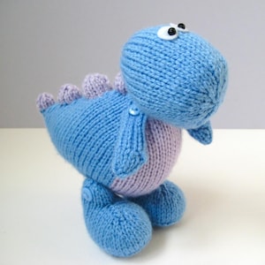 Puede incluir: Un peluche de dinosaurio tejido a mano en azul y morado claro. La criatura presenta una cabeza redondeada con dos ojos de plástico en blanco y negro, una barriga morada clara y una hilera de pequeñas púas moradas claras a lo largo de la espalda. Pequeños botones azules sujetan los brazos y las piernas al cuerpo.
