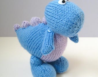 May include: A hand-knitted blue and light purple dinosaur plush toy. The creature features a rounded head with two black and white plastic eyes, a light purple belly, and a row of small light purple spikes along its back. Small blue buttons attach the arms and legs to the body.