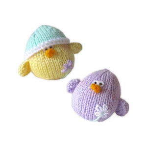 May include: Two knitted bird-shaped toys. One is yellow with a light blue and white hat, orange beak, and a purple flower. The other is purple with an orange beak and a white flower. Both have small wings.