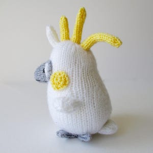 May include: A hand-knitted cockatoo plush toy featuring a white body, a bright yellow crest on its head, a yellow circular patch on each cheek, and a grey beak and feet. The bird is depicted in a side profile view with a soft, textured yarn finish.