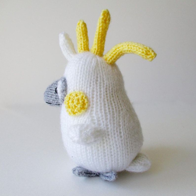 May include: A hand-knitted cockatoo plush toy featuring a white body, a bright yellow crest on its head, a yellow circular patch on each cheek, and a grey beak and feet. The bird is depicted in a side profile view with a soft, textured yarn finish.