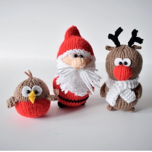 May include: Three knitted figurines including a round brown and red bird with a yellow beak, a Santa Claus figure in a red suit with a white beard, and a brown reindeer with black antlers, a red nose, and a white scarf against a white background.