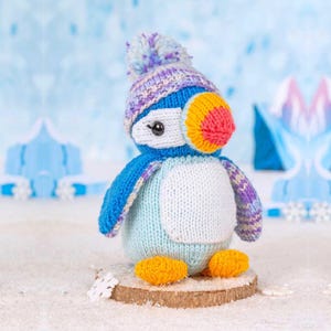May include: A knitted puffin toy featuring a light blue body, a white belly, and dark blue wings. It wears a purple, white, and blue striped hat with a matching pom-pom on top. The puffin has a bright orange and yellow beak and orange feet.
