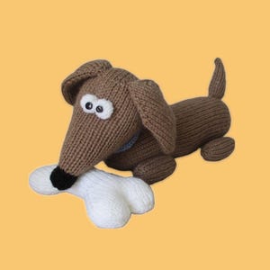 Könnte beinhalten: Ein braunes, gestricktes Dackel-Spielzeug mit großen Schlappohren, weißen Augen und einer schwarzen Nase, das einen weißen Knochen hält. Der Hund hat ein blaues Halsband und einen kleinen Schwanz. Das Spielzeug ist vor gelbem Hintergrund.