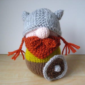 May include: A knitted gnome figurine wearing a grey horned helmet, a pale pink nose, and a thick orange beard. The body features horizontal stripes of yellow and brown yarn. The gnome has thin orange arms with fringed ends and holds a round shield.