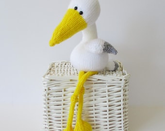 May include: A knitted white bird toy with a long yellow beak, small black eyes, and grey wing tips. The bird has long, dangling yellow legs with webbed feet, and it is positioned on top of a white woven wicker basket with a rectangular shape.