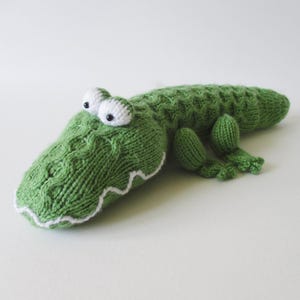 Puede incluir: Un juguete de cocodrilo tejido en color verde con un patrón de trenzas texturizado en el lomo, ojos tejidos en blanco con pupilas negras y un detalle de boca con costura en zigzag blanca. El peluche tiene cuatro patas cortas y una cola larga.