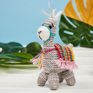 May include: A hand-knitted grey llama figurine featuring a white muzzle, a teal halter, and a colorful woven saddle blanket with pink fringe. The llama has upright ears, embroidered black eyes, and a small tail, standing on four sturdy legs.