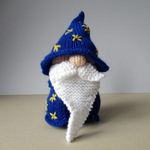 May include: A knitted gnome figurine with a blue wizard hat adorned with yellow stars. The gnome has a white beard, a tan nose, and is wearing a blue robe. The handmade craft is a whimsical decorative item.