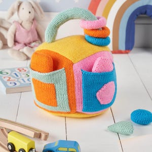 May include: A colorful knitted toy with a handle, shaped like a house with different colored geometric shapes and pockets. The toy has a set of colorful rings that can be stacked on the handle.