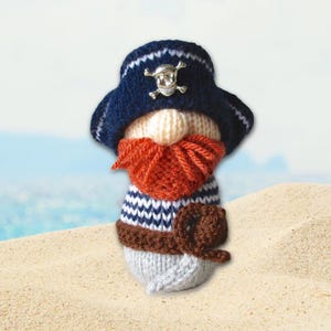 May include: A handcrafted pirate gnome doll with a blue hat featuring a skull and crossbones emblem. The gnome has a cream-colored beard, an orange mustache, and a striped shirt. It is holding a brown hook and is set against a beach background.