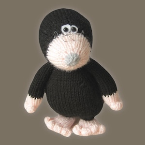 May include: A knitted mole toy, predominantly black with a light pink face, hands, and feet. It features small wire-rimmed spectacles and a grey nose. The toy has a rounded body and small limbs, giving it a charming, whimsical appearance.