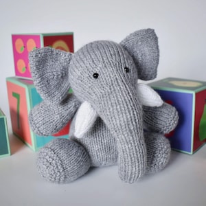 May include: A grey knitted elephant plush toy with large ears, a long trunk, and two white tusks. The elephant has small black bead eyes and is constructed from a textured yarn material. It is positioned in front of colourful toy blocks.