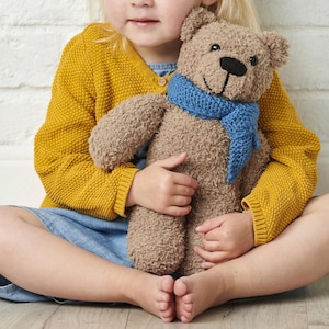 May include: A brown teddy bear with a blue scarf. The bear has a black nose and smiling mouth. The bear is being held by a person wearing a yellow cardigan and a blue dress.