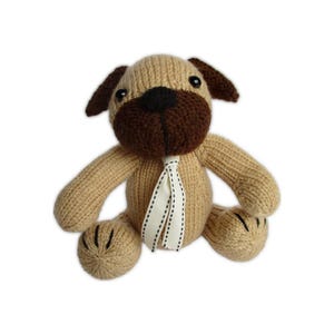 May include: A knitted stuffed animal shaped like a dog with a tan body, dark brown ears, and a dark brown muzzle. The dog has black plastic eyes and a white ribbon tied around its neck with a thin black stitched line running down the center.