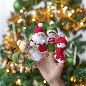 May include: A hand displaying five knitted Christmas finger puppets. The puppets include Santa Claus, an elf, a snowman, an angel, and a reindeer. The puppets are red, green, white, and beige, with white pom-poms and festive details.