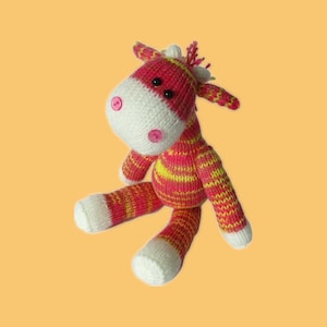 May include: A handmade knitted toy horse in shades of pink, yellow, and white. The horse has a white face with pink button eyes and pink button accents. The body is striped with pink and yellow, and the legs and ears are also striped.
