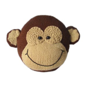 May include: A knitted monkey face pillow in shades of brown and tan. The monkey has large, tan eyes, a tan muzzle, and a smiling mouth. The ears are tan and brown. The pillow is round and appears to be handmade.