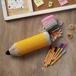 May include: A yellow knitted pencil case with a black tip, gray eraser, and pink top, filled with pencils. The case is open, revealing the pencils inside. Colorful pens and fruit-shaped erasers are scattered around.