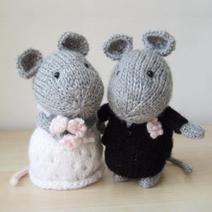 Puede incluir: Dos figuras de ratones tejidas a mano en color gris. Un ratón lleva un vestido blanco texturizado y sostiene un pequeño ramo de flores de color rosa claro. El otro ratón lleva una chaqueta negra tejida con una flor rosa claro en el pecho.