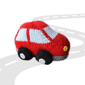 Pode incluir: Um carro de brinquedo vermelho tricotado à mão, com janelas azuis claras e rodas pretas. O carro tem um botão amarelo como farol e está sobre um fundo branco com um desenho de estrada. Um brinquedo macio para crianças.