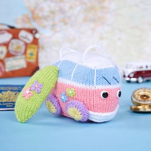 May include: A knitted toy bus in pastel shades of pink, blue, and white. It has black button eyes, a green surfboard with flower details, and purple and yellow wheels. A cute, handmade travel-themed item.