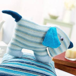 Op de afbeelding: Een gebreid honden speelgoed in blauw-wit gestreept ontwerp met blauwe oren en een zwarte staart. Het speelgoed zit op een blauw-wit gestreept gebreid kussen.