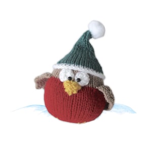 Könnte beinhalten: Gestrickte Rotkehlchen-Vogel-Figur mit roter Brust, braunen Flügeln und einer grünen Mütze mit weißem Bommel. Der Vogel hat weiße Augen, einen gelben Schnabel und sitzt auf einer hellblauen Oberfläche. Ideal für die Weihnachtsdekoration.