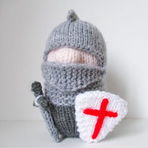 May include: A knitted toy shaped like a knight wearing a grey helmet and armour. The figure has a pale pink face visible through the helmet opening, holds a grey sword in one hand, and carries a white shield featuring a bright red cross emblem.
