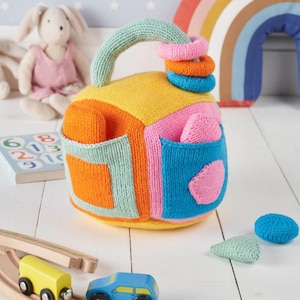 May include: A brightly coloured knitted baby toy with a handle and three stacking rings. The toy has a patchwork design with orange, yellow, pink, and blue sections. Two pockets and two additional knitted shapes are also visible. A plush bunny and wooden toy train are in the background.