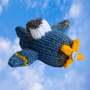 May include: A knitted blue airplane toy with yellow and gray accents. The wings have yellow stripes, and the propeller is yellow with a blue button in the center. The plane's top is white and gray.