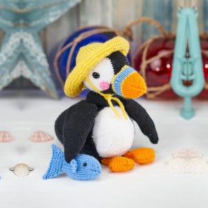May include: A handcrafted knit puffin toy featuring a black and white body, a bright yellow sun hat with a chin strap, an orange and blue beak, and pink cheeks. The puffin is holding a small blue knit fish in one wing and has orange feet.