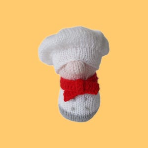 Puede incluir: Un gnomo de chef tejido con un gorro de chef blanco y una pajarita roja. El cuerpo del gnomo es blanco con una base gris y detalles plateados. El fondo es de color amarillo-naranja sólido.