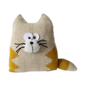 Op de afbeelding: Een gebreid katvormig kussen in crème en mosterdgeel. De kat heeft grote witte ogen, een witte neus en zwarte snorharen. De staart is gestreept met crème en geel. Een decoratief item.