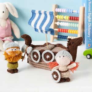 Puede incluir: Un barco vikingo de juguete tejido con una vela a rayas azules y blancas, acompañado de dos figuras vikingas tejidas. El barco es marrón con detalles circulares, y las figuras tienen sombreros, barbas y escudos.