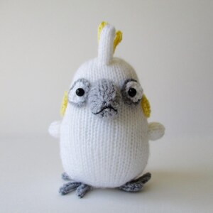 May include: A white knitted cockatoo plush toy featuring a grey beak and eye patches, a yellow crest on top of its head, yellow cheek patches, and small grey feet. The bird has two black bead eyes and a rounded, soft-textured body structure.