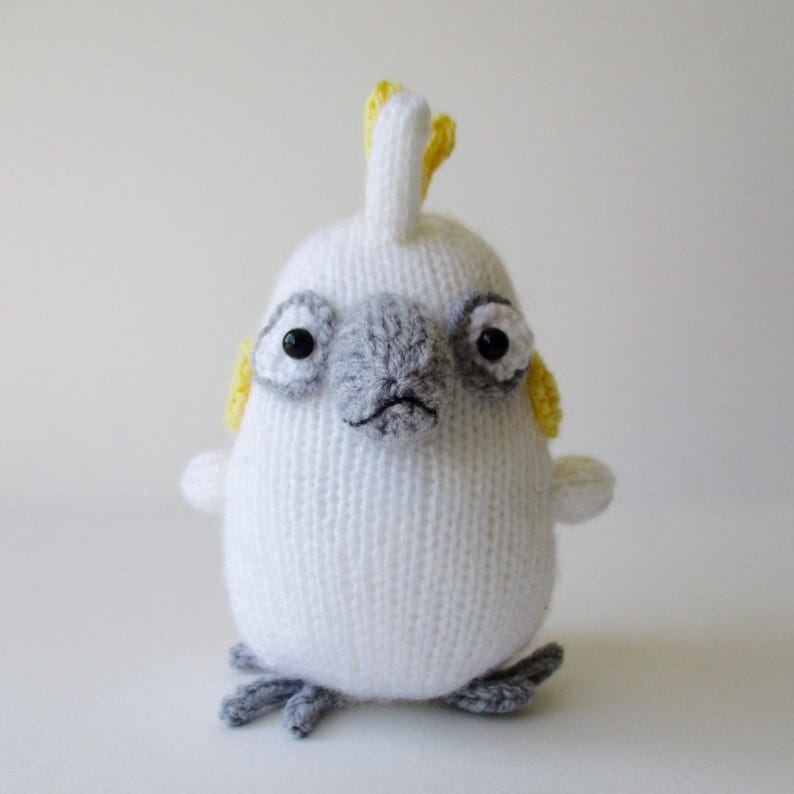 May include: A white knitted cockatoo plush toy featuring a grey beak and eye patches, a yellow crest on top of its head, yellow cheek patches, and small grey feet. The bird has two black bead eyes and a rounded, soft-textured body structure.