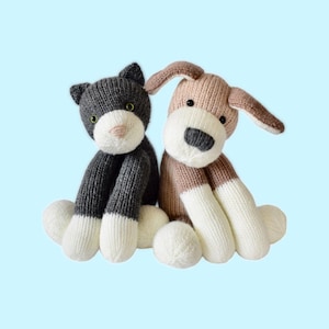 Op de afbeelding: Twee gebreide knuffels: een grijze en witte kat en een bruine en witte hond. De kat heeft groene ogen en een roze neus. De hond heeft een zwarte neus en slappe oren. Beide zitten op een lichtblauwe achtergrond.