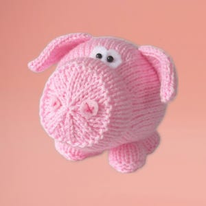 May include: A handmade pink knitted pig toy with a round body, large ears, and small black eyes. The pig has a detailed snout with a small pink cross-stitch detail. The toy is made of soft yarn and is suitable for children.