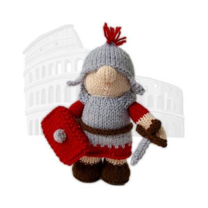 Puede incluir: Muñeco de soldado romano tejido, con escudo rojo y espada plateada. El muñeco lleva casco y túnica grises, detalles rojos y botas marrones. El Coliseo es visible en el fondo.