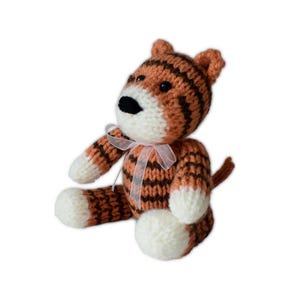 May include: A handmade knitted tiger toy with orange and brown stripes. The tiger has a white muzzle, paws, and a small pink ribbon tied around its neck. The toy is sitting upright.