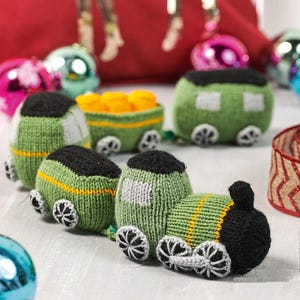 May include: Hand-knitted toy train set with a green and black color scheme. The train cars have white and yellow stripes, and the wheels are gray and white. The train cars are filled with yellow objects. The background includes Christmas ornaments.