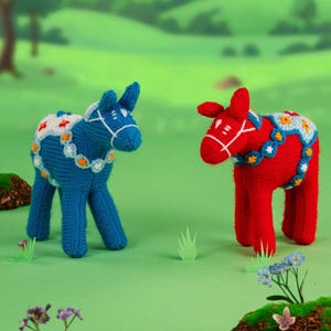 May include: Two knitted toy horses, one blue and one red, stand on a green surface. The blue horse has a white and orange floral design on its back. The red horse has a similar design. Both have white bridle details.