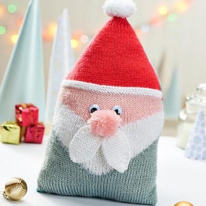 以下が含まれることがあります： 編み物で作られたサンタクロース型のギフトバッグ。赤い帽子に白いポンポン、ピンクと白の顔、動く目、ピンクのポンポンの鼻、白いひげのディテールが特徴です。バッグの底は緑色です。