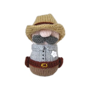 Può includere: Uno gnomo fatto a mano, vestito da cowboy. Lo gnomo indossa un cappello da cowboy color camoscio, una camicia grigia con una stella da sceriffo, una cintura marrone con fibbia dorata e una base lavorata a maglia marrone. Ha un naso rosa e baffi grigi.