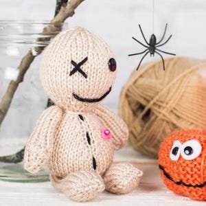 May include: A beige knitted doll with a round head, one black button eye, one black stitched X eye, and a black stitched smile. The doll features a small pink bead on its chest and black vertical stitching down the centre of its torso.