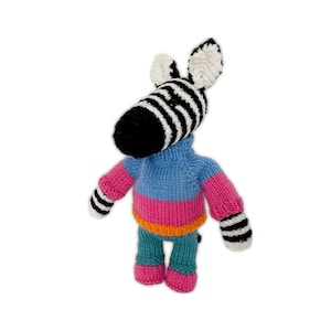 May include: A hand-knitted zebra plush toy featuring a black and white striped head and limbs. The zebra wears a sweater with a light blue upper section, a horizontal pink stripe, and an orange hem, paired with teal pants and pink feet.